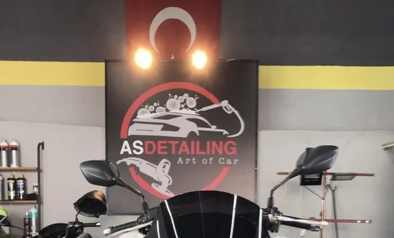 Atölyede iki spot ışığın altında asılı AS Detailing tabelası, önünde bir motosiklet